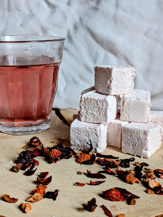 Verre d'infusion rouge rubis à côté d'une pyramide de guimauves artisanales roses sur un rondin de bois, ambiance goûter gourmand et naturel.