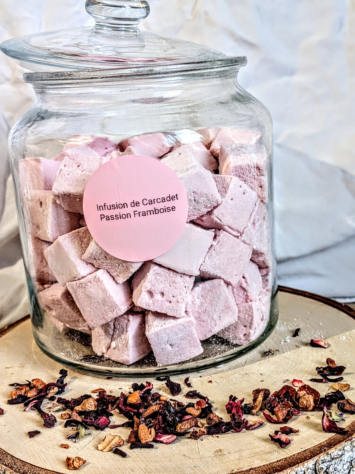 Bocal de guimauves artisanales roses Les Tabliers Enchantés parfumées à l'infusion de Carcadet, Passion et Framboise, posé sur un rondin de bois avec des éclats de fruits séchés. Artisanat Orléans