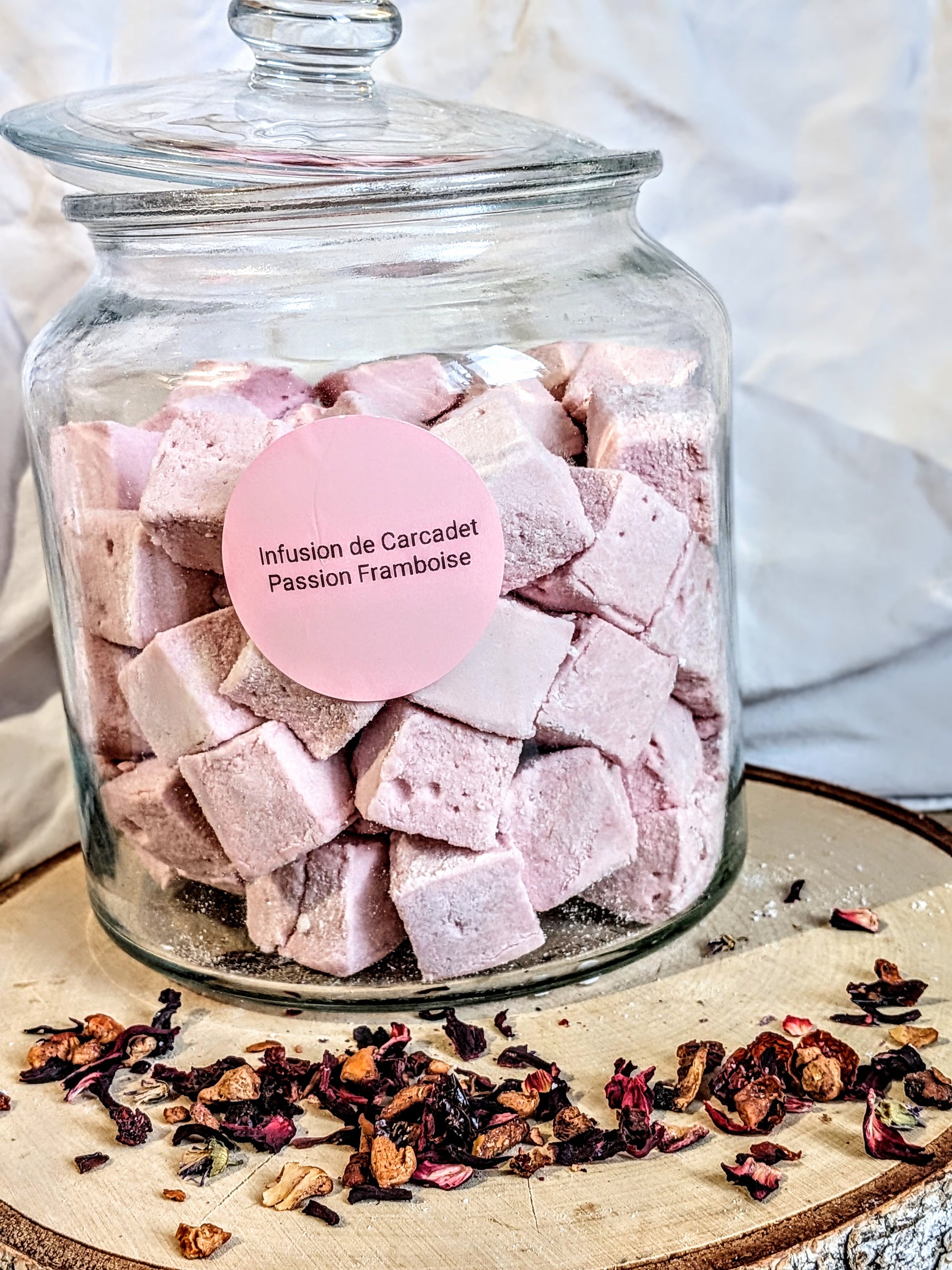Bocal de guimauves artisanales roses Les Tabliers Enchantés parfumées à l'infusion de Carcadet, Passion et Framboise, posé sur un rondin de bois avec des éclats de fruits séchés. Artisanat Orléans