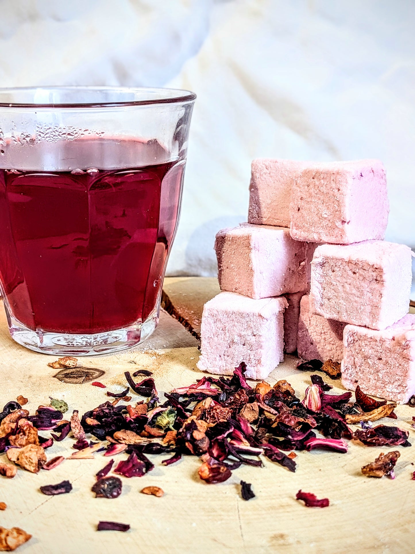 Un verre d'infusion rouge intense à côté d'une pile de guimauves à l'infusion carcadet passion framboise, entourés de fleurs d'hibiscus et de morceaux de fruits exotiques séchés